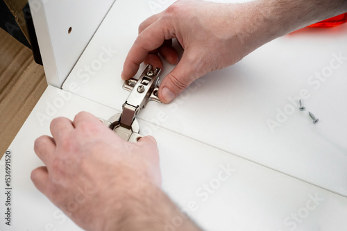Hands installing a metal hinge on a white cabinet door with two screws nearby