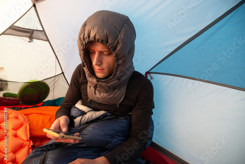 Man in a sleeping bag, wearing a down sleeping hood, using smartphone inside a tent during winter camping