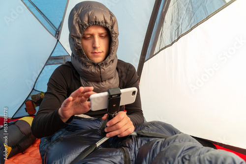 Man in a sleeping bag, wearing a down sleeping hood, using smartphone inside a tent during winter camping