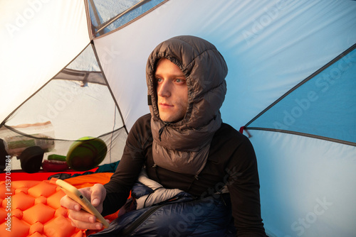 Man in a sleeping bag, wearing a down sleeping hood, using smartphone inside a tent during winter camping