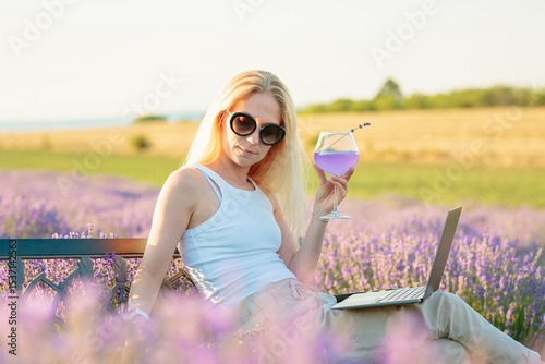 Lavender field. Woman sitting on bench among lavender flowers and working on laptop computer with glass of summer refreshing cocktail, relaxing, resting, enjoying nature. Mental health concept