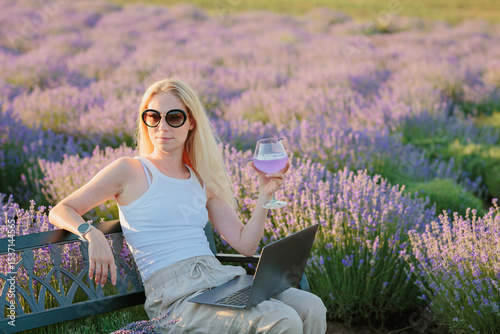 Lavender field. Woman sitting on bench among lavender flowers and working on laptop computer with glass of summer refreshing cocktail, relaxing, resting, enjoying nature. Mental health concept
