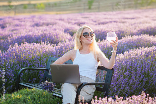 Lavender field. Woman sitting on bench among lavender flowers and working on laptop computer with glass of summer refreshing cocktail, relaxing, resting, enjoying nature. Mental health concept