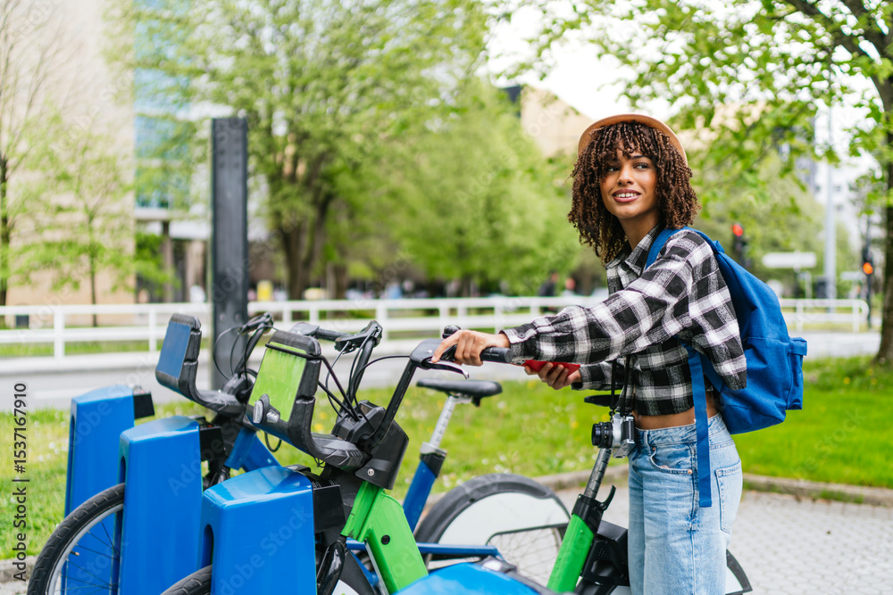 Obraz premium Young latin student renting a bike sharing service using her smartphone app, sustainable mobility concept