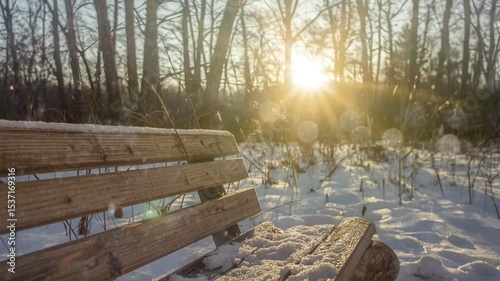 Close-up of a wooden bench lightly covered with snow in a quiet winter forest. Sunlight filters through bare trees, creating a soft bokeh effect in the background.