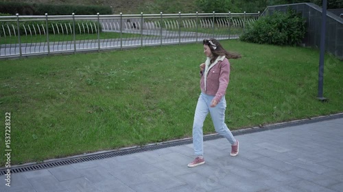 Youthful female dancing freely, listening to music with wireless headphones, moving energetically through city park, embodying carefree urban lifestyle and personal expression