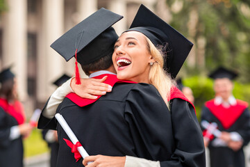 Cheerful young blonde lady hugging her black boyfriend while graduation ceremony at university campus. Two happy multiracial students greeting each other with successful graduation, closeup