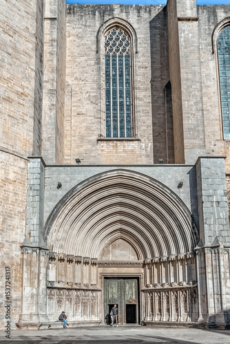Gothic entrance of Girona Cathedral with ornate arched portal and tall stained glass window. Visitors at the doorway highlight the scale of the medieval structure. Girona, Spain - November 28, 2021