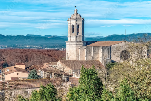 Ancient Girona Cathedral with its bell tower rises above medieval stone buildings and terracotta roofs. Trees and distant mountains form a picturesque Catalan backdrop