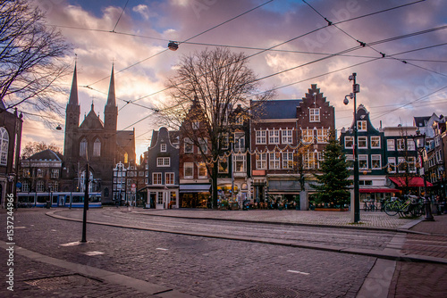 Amsterdam, Netherlands. A city square with cobblestone streets and traditional Dutch buildings adorned with holiday lights. A tram is making its way through the square along the tracks.