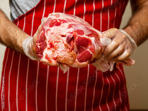 Fresh juicy leg of lamb with bone in a butcher hands. Red and white stripe apron background. Selective focus. High quality and price meat product. Agriculture and food industry.