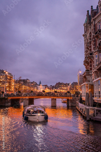 Amsterdam. The city glows, adorned with various lights and garlands. A boat glides through the canal, with the shimmering reflections of the lights on the water creating a festive atmosphere.