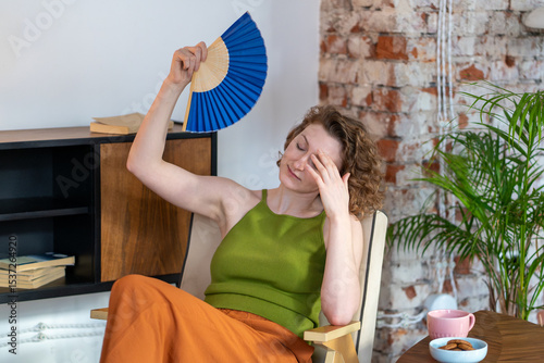 Heated fatigued woman suffers from lack of air conditioning in home. Tired overheated female waves hand fan to find relief from high temperature feels headache. Discomfort, weakness on sweltering day.
