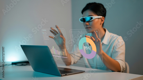 Woman using augmented reality glasses interacting with holographic data near laptop on desk