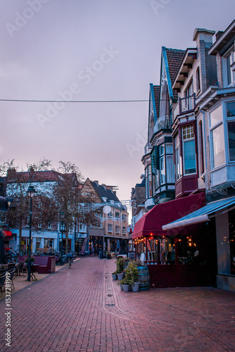 A narrow street in Amsterdam, lined with beautiful, colorful houses, various cafes and pubs, with bicycles parked along the way.