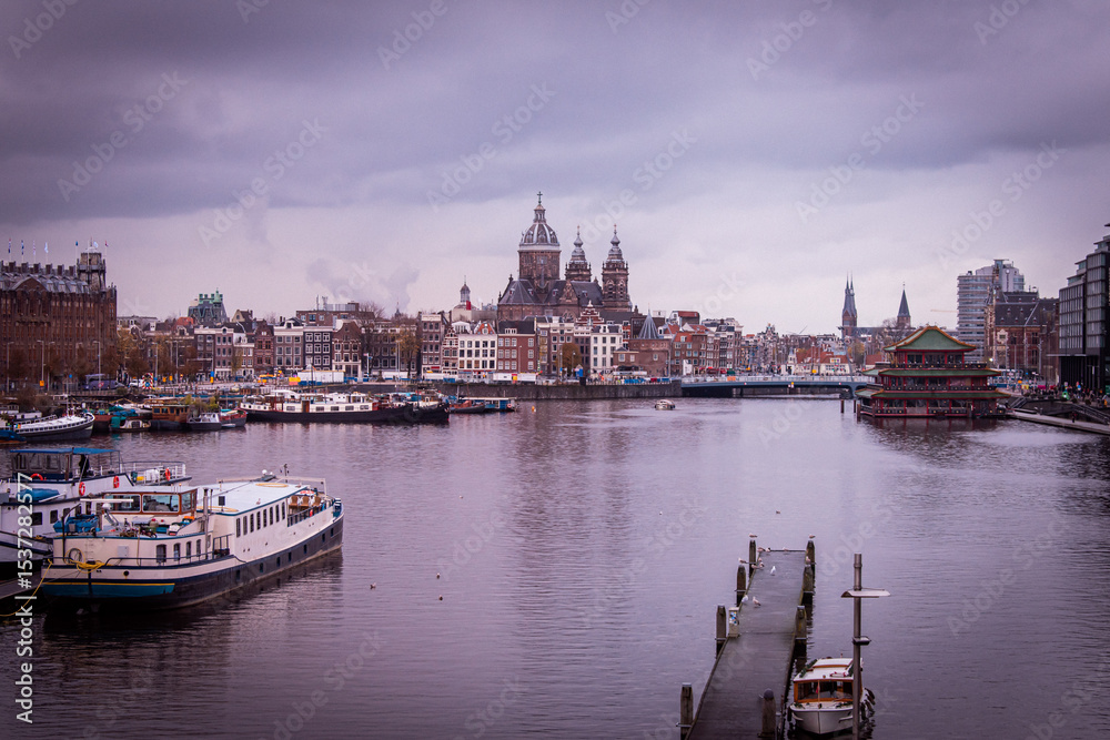 Fototapeta premium Amsterdam, Netherlands. The features boats docked along the canal. In the background, the Basilica of Saint Nicholas with multiple spires and domes. 
