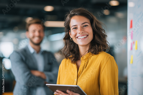 Mujer sonriente con tableta en una oficina moderna, con un hombre desenfocado al fondo, ambiente laboral positivo. Liderazgo femenino, confianza radiante, espíritu colaborativo, innovación continua.