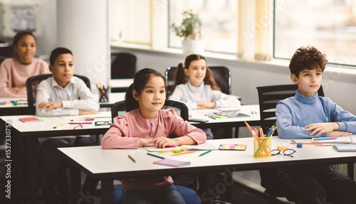 Back To School. Portrait of focused multicultiral pupils sitting with folded arms at desks with social distance, listening to teacher. Diverse junior students working diligently and learning new stuff