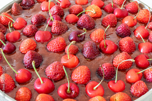 A freshly homemade round cake topped with coulis sauce and whole strawberries and cherries with stems, placed in a metal baking pan. Macrophotograph detailed closeup oblique overhead view.