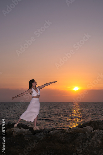 Fit girl strikes a dancer pose on a cliff at sunset in Ibiza