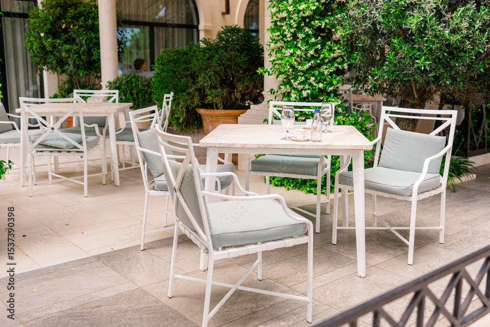 Obraz premium Elegant outdoor cafe terrace with white furniture and lush greenery. Relaxation, summer lifestyle, and modern hospitality.