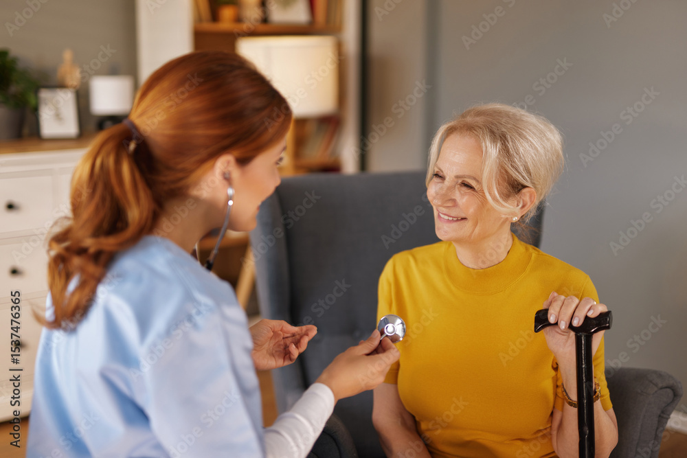 Fototapeta premium A young woman with red hair, wearing a blue uniform, uses a stethoscope on a smiling senior woman with short blond hair in a yellow shirt. The senior woman is sitting in a chair with a cane.
