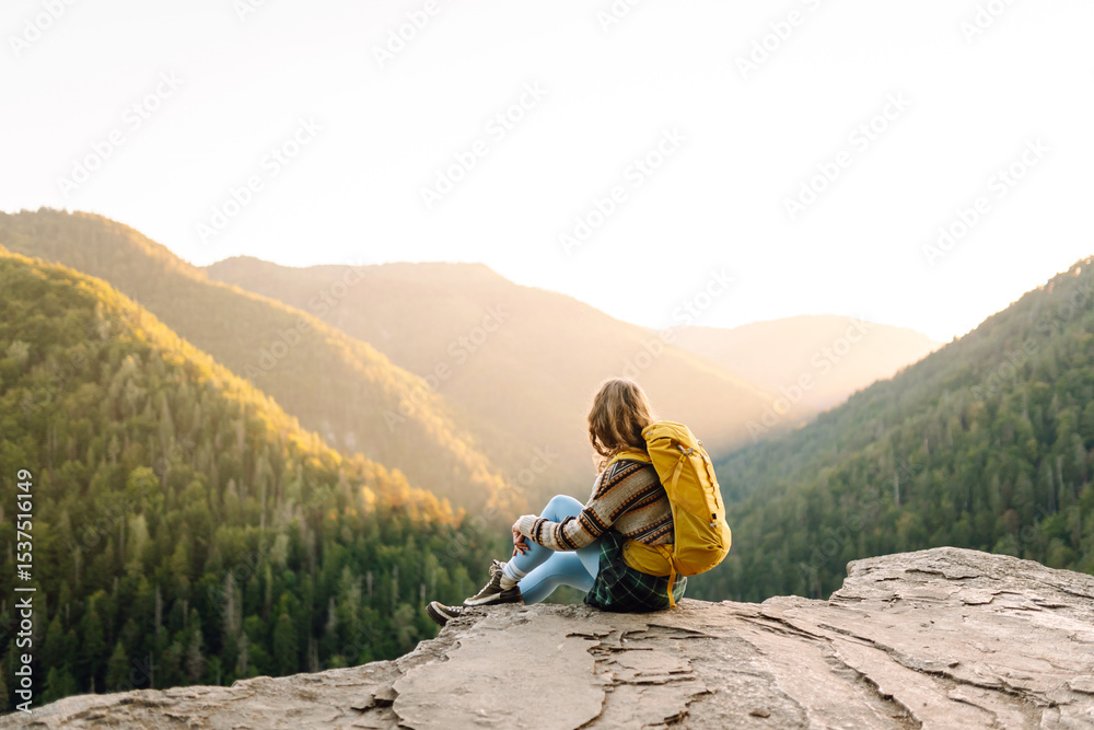 Fototapeta premium Beautiful woman hiker feels freedom standing on rocky ledge enjoying sunset in mountains. Young female hiker looks at mountain landscape on cliff. Hiking, adventure and nature concept.