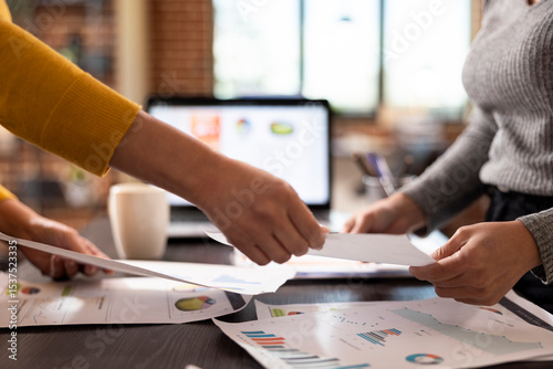 Closeup of hands of female accountant exchanging documents, collaborating at office desk in analyzing company expenses report. Selective focus of individuals holding papers over the table.