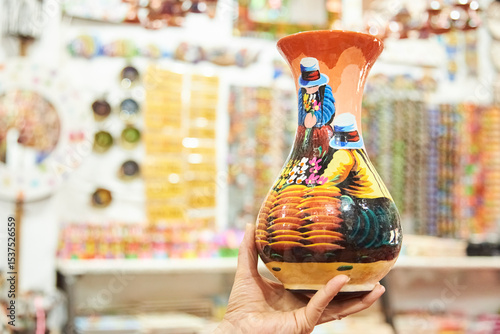 Female hand holding a handcrafted ceramic vase painted with a colorful traditional scene, in Raquira, Colombia, town known for its handicrafts and pottery.