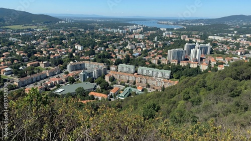 Aerial view of a cityscape with residential buildings, greenery, and distant water under a clear blue sky