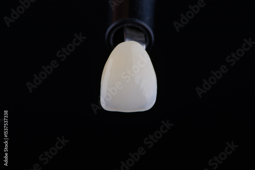 Close up shot ultra thin dental zirconium crown is held with tweezers with black background.