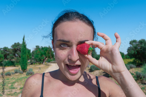 Mujer deportista con una fresa en el ojo y la boca abierta, un día soleado con cielo azul en el parque y enfoque selectivo.