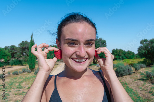 Mujer deportista con dos fresas en las orejas como pendientes y una sonrisa, un día soleado con cielo azul en el parque y enfoque selectivo.