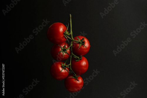 Cluster of vine tomatoes hanging against a dark background. Ripe red tomatoes with natural shine and texture. Minimalist organic food concept.

