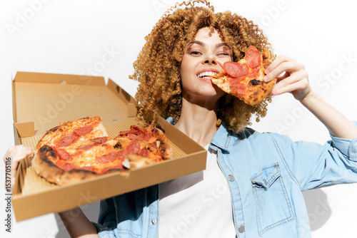 Young woman enjoying a delicious pepperoni pizza slice, showcasing joy and excitement against a clean, white background with warm tones