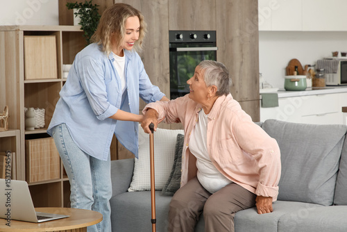 Tableau sur toile Young woman helping her grandmother with stick to stand up from sofa at home