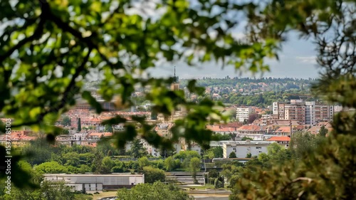 Central European city skyline framed by green trees. Features historic tower, modern buildings, residential areas under a partly cloudy sky. Ideal for travel & urban themes.