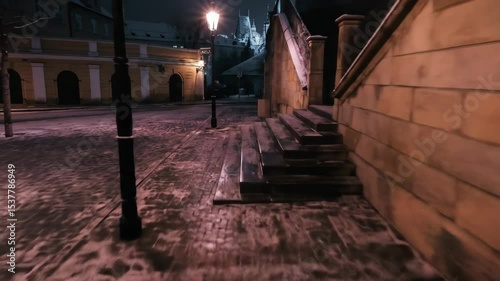 Low angle tracking shot over snowy cobblestone street at night. Illuminated by warm streetlights, the historic buildings and empty path evoke a quiet, mysterious European winter.