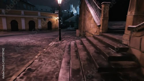 Low angle tracking shot over snowy cobblestone street at night. Illuminated by warm streetlights, the historic buildings and empty path evoke a quiet, mysterious European winter.