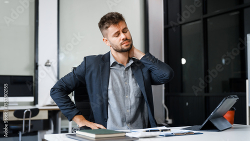 A stressed Caucasian businessman in a suit sit indoors at an office desk, suffering from office syndrome symptoms including headache, neck pain, and wrist pain due to work-related strain.