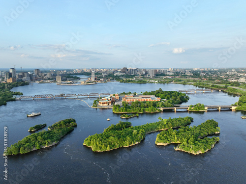 Aerial view of lush green islands nestled within the wide Ottawa River, with bridges connecting them and the city skyline of Ottawa visible in the background under a clear sky