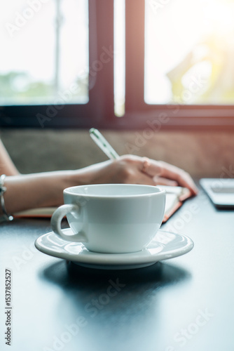 A serene morning setup featuring a white coffee cup and a person writing in a notebook by a sunlit window.