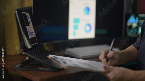A person is writing on a piece of paper in front of a computer monitor. The person is using a pen and the paper is on a clipboard. The computer monitor shows a graph