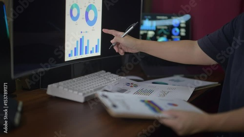 A person is sitting at a desk with a computer monitor and a keyboard. They are holding a clipboard and a pen, and are looking at a graph on the computer screen. The graph appears to be a pie chart