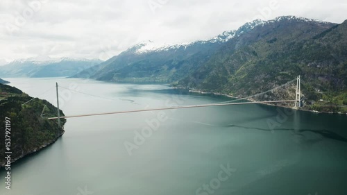 Hardanger Bridge arches over wide fjord flanked by imposing mountains with snow caps. Long bridge provides striking contrast in serene landscape