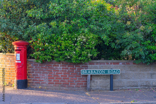 The iconic red postbox stands next to a street sign on Brabazon Road in Norwich, UK. Vintage British postal pillar box against a brick wall and trees