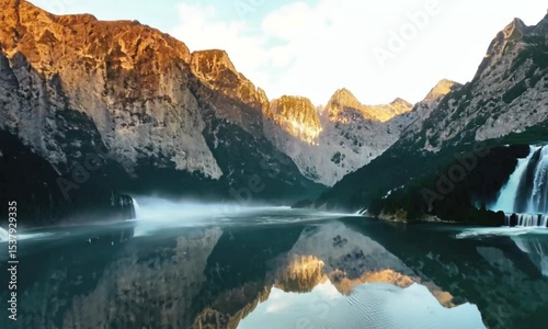 A beautiful mountain range with a waterfall in the foreground. The water is calm and the sky is clear