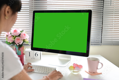 A woman is sitting at a desk with a green computer monitor in front of her. The monitor is blank, and the woman is typing on a keyboard. The room has a floral arrangement on the desk and a pink cup
