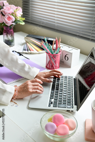 A woman is typing on a laptop in front of a white desk. The desk is cluttered with various items such as a bowl of pink macarons, a vase with flowers, a cup, a book