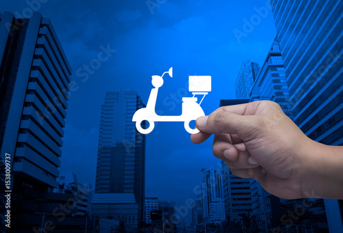 Hand holding motorcycle flat icon in fingers over modern office city tower and skyscraper, Business delivery service concept